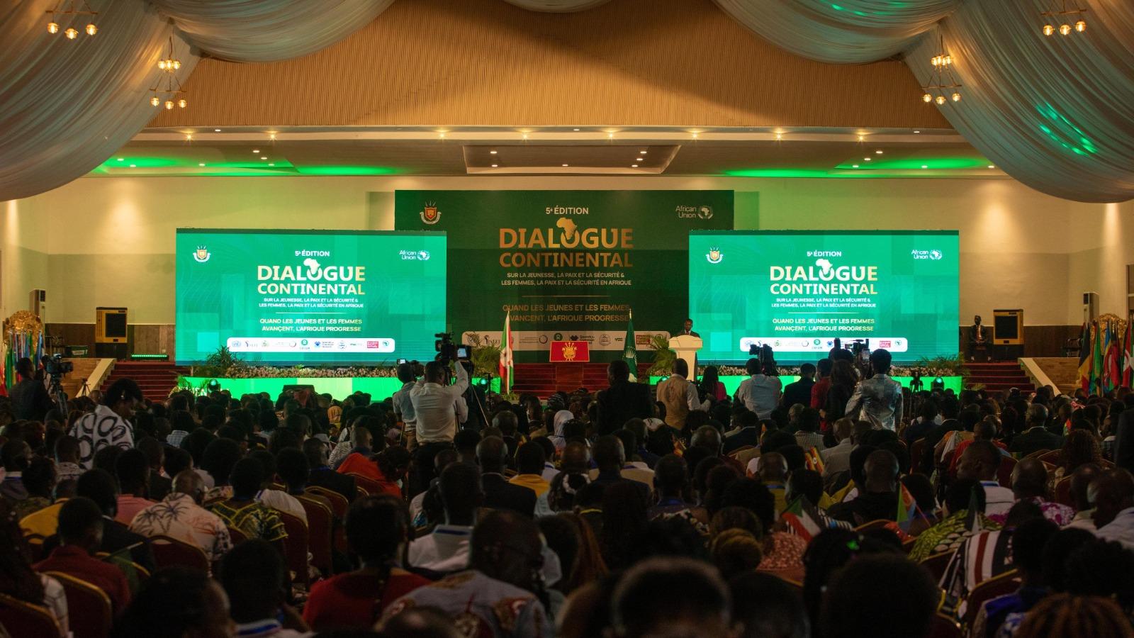 A packed conference hall with draped ceilings, green-lit screens reading “Dialogue Continental,” a stage, flags, and an attentive audience.