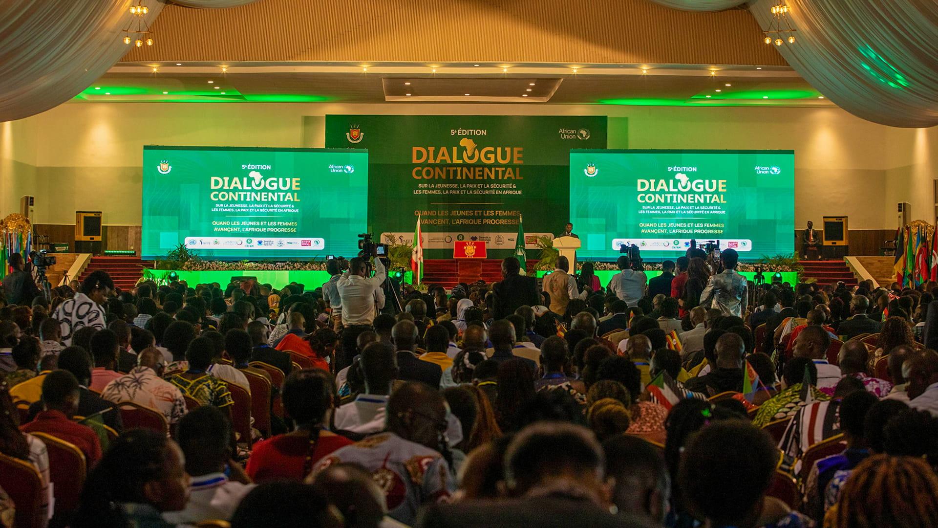 A packed conference hall with draped ceilings, green-lit screens reading “Dialogue Continental,” a stage, flags, and an attentive audience.