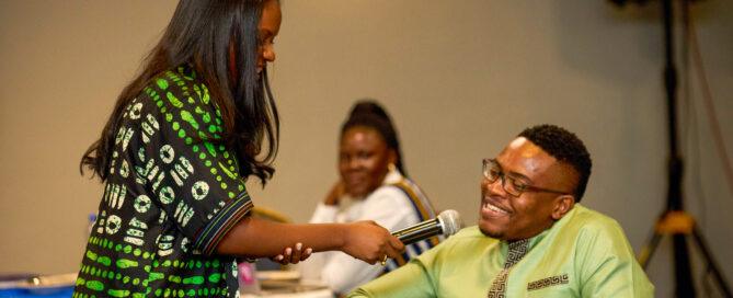 A woman in a green and black patterned dress holds a microphone to a smiling man seated in a green shirt. A woman in the background watches attentively