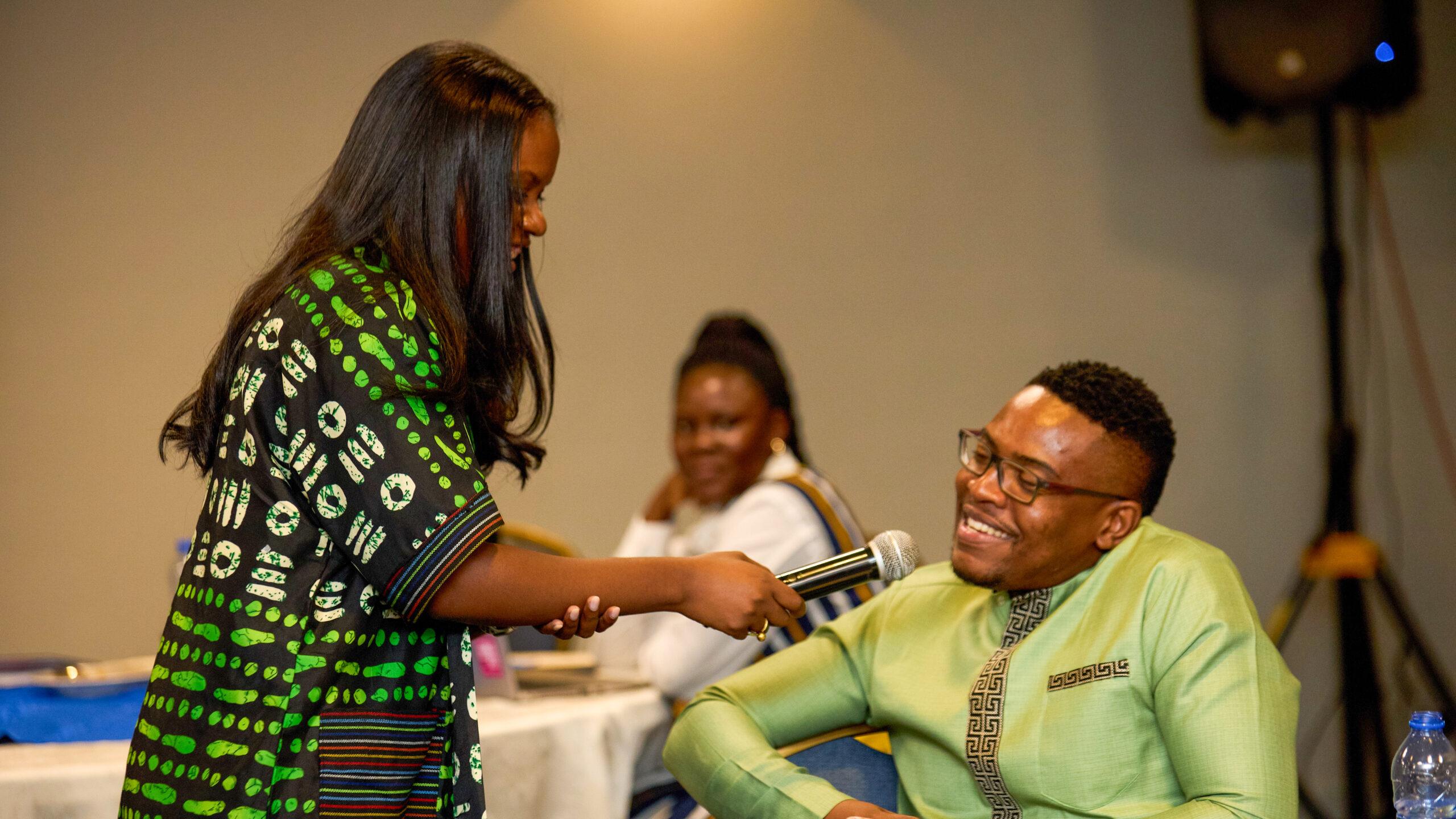 A woman in a green and black patterned dress holds a microphone to a smiling man seated in a green shirt. A woman in the background watches attentively