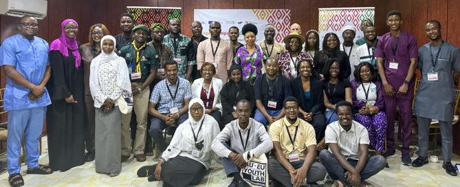 A diverse group of around 30 people pose together in a room with wood-paneled walls. They wear name tags, smiling, conveying a sense of unity and collaboration.