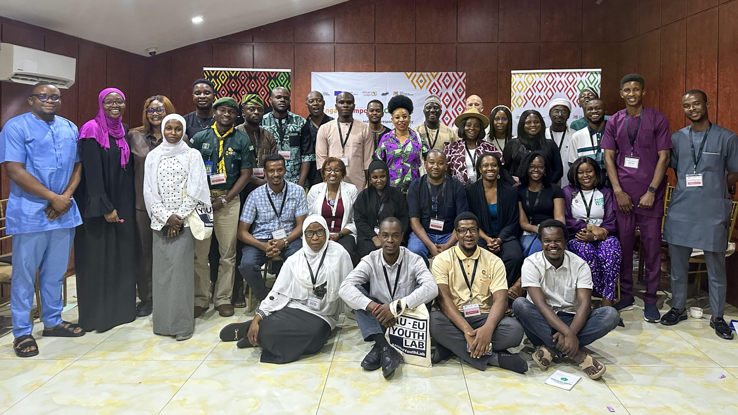 A diverse group of around 30 people pose together in a room with wood-paneled walls. They wear name tags, smiling, conveying a sense of unity and collaboration.