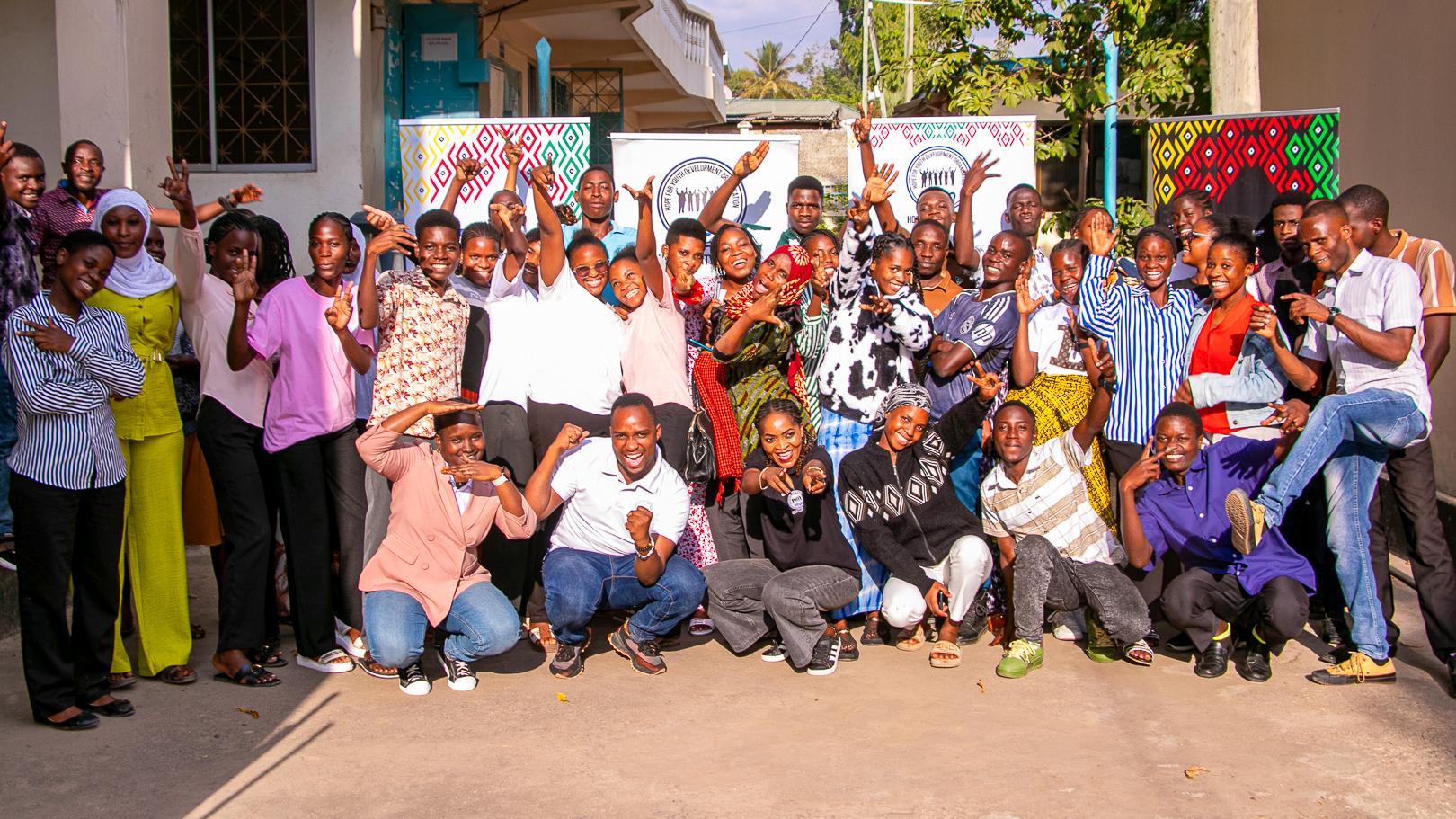 Large, joyful group of diverse people posing outdoors, raising hands and smiling beside colorful patterned posters in bright sunlight.