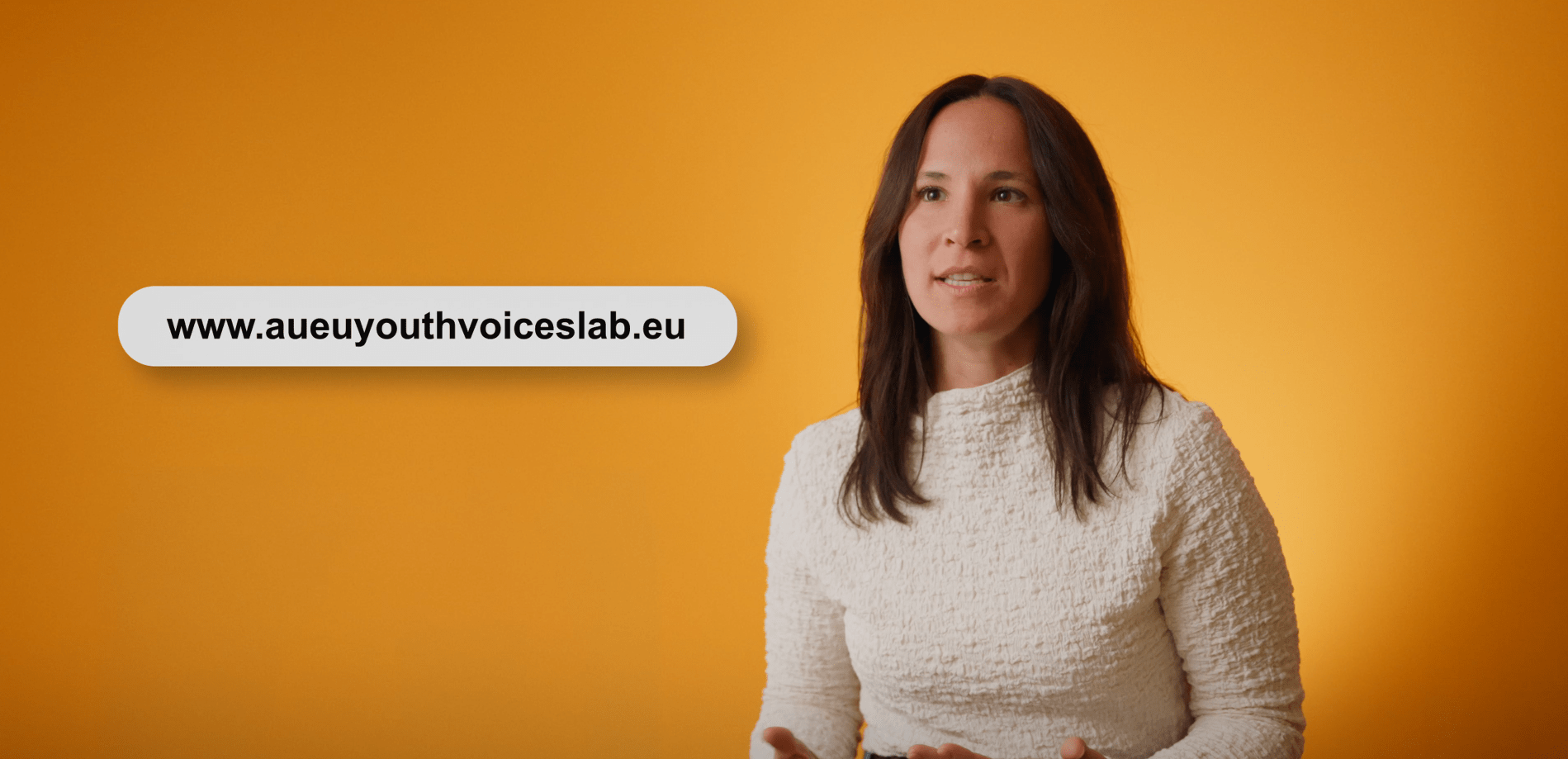 A woman in a white top speaks in front of an orange background. A website URL, www.aueyouthvoiceslab.eu, is displayed in a rounded box to the left of her.