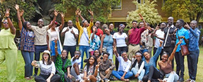 A diverse group of young adults joyfully poses outdoors on a lawn, with trees and a building in the background. They exude happiness and camaraderie.