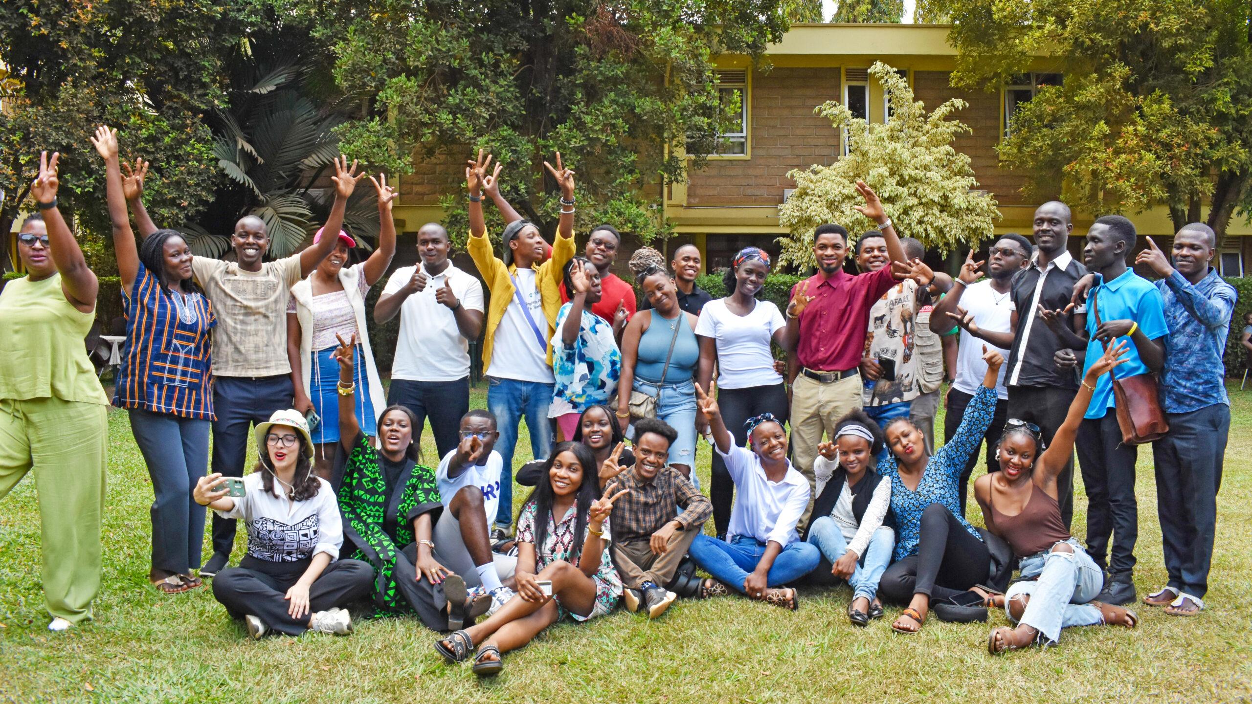 A diverse group of young adults joyfully poses outdoors on a lawn, with trees and a building in the background. They exude happiness and camaraderie.