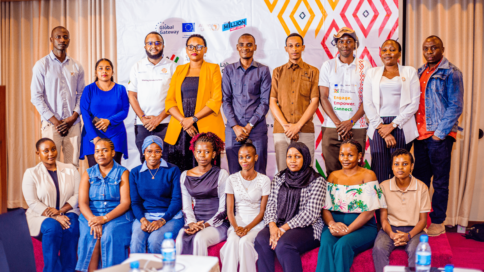 Seventeen people pose for a group photo indoors in front of a branded backdrop, with some standing and others seated, dressed in casual and business attire.