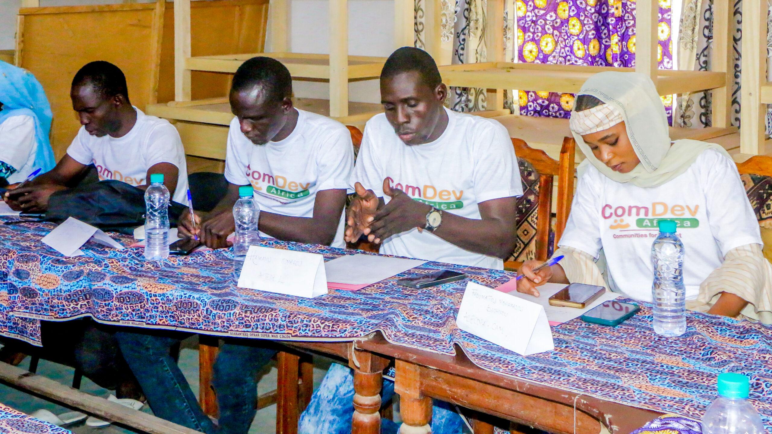 Five individuals are seated at a table covered with a patterned cloth, wearing "ComDev" T-shirts, attentively writing and discussing at an event.